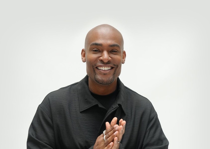 Paul C Brunson smiling towards the camera in front of a white backdrop, wearing a black tshirt and jacketx